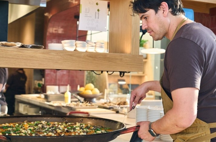 Chef tending a large paella pan at Picala's open kitchen, wearing an apron and focused on the dish.