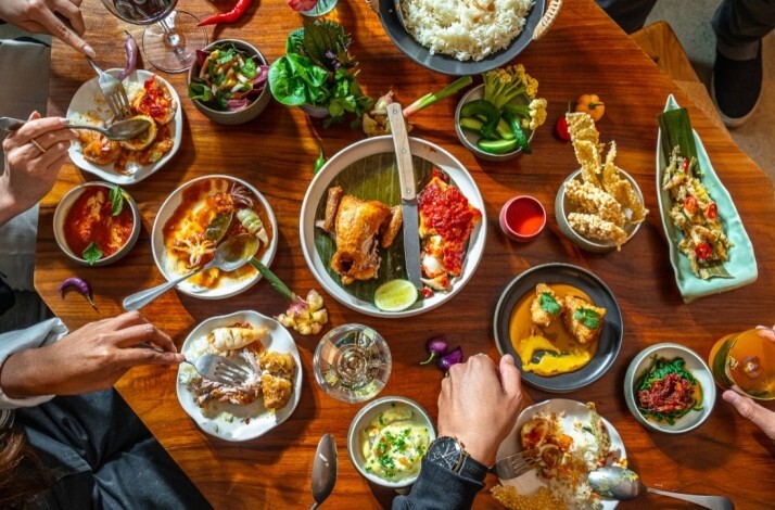 Overhead view of a wooden table filled with Indonesian Padang-style dishes, rice, curries, sambal, and shared plates as diners reach in at Ma.dé.