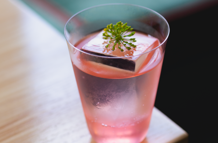 Pink cocktail in a clear glass with a large ice cube and a small herb garnish, served at Firstborn.
