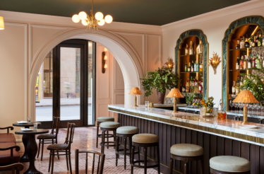 Modern French bistro bar with marble counter, green-cushioned stools, arched doorway, and warm lighting.