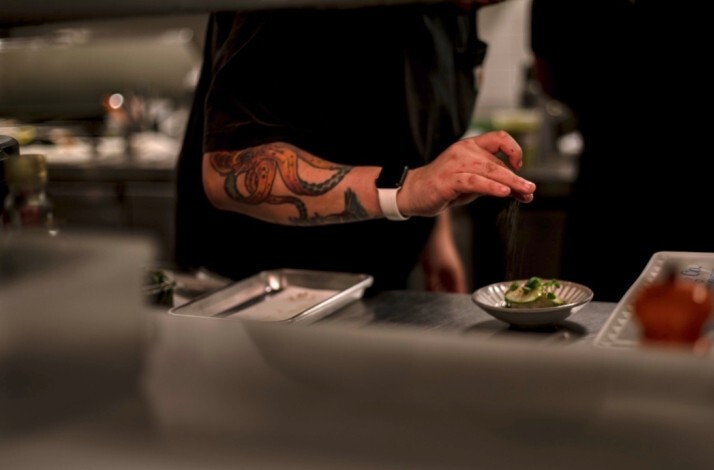 Chef with octopus forearm tattoo finishing a plated dish with a pinch of seasoning at Zitz Sum.