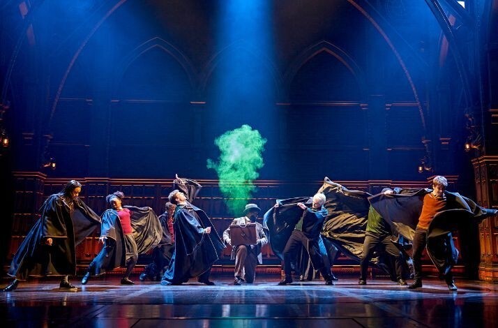 Ensemble cast in dark robes performs a spell as green smoke rises center stage under dramatic blue lighting at a Harry Potter and the Cursed Child showing.