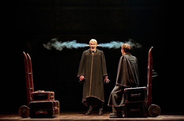 Two cloaked men face each other between wheeled trunks as a burst of smoke shoots from one man’s ears in a dark, spotlighted scene at a Harry Potter and the Cursed Child showing.