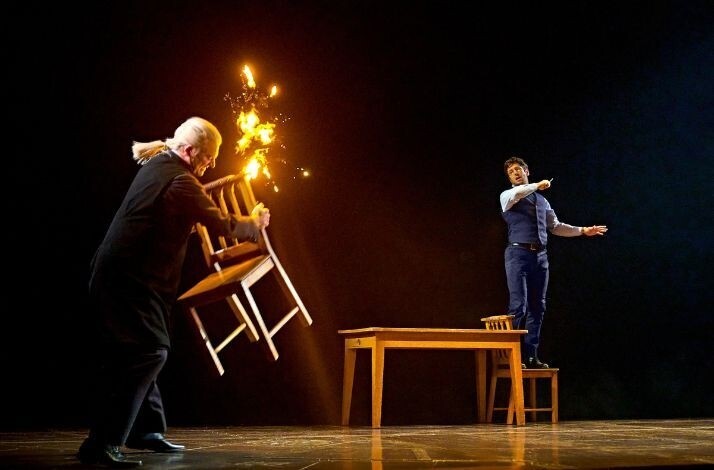 Onstage magic moment where a chair bursts into flames as one performer lunges forward and another stands on a chair by a table, reacting.