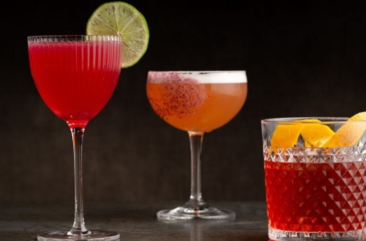 Trio of artisan cocktails at Nightbird, featuring vibrant red, amber, and dark ruby drinks garnished with lime, spiced rim, and orange peel.