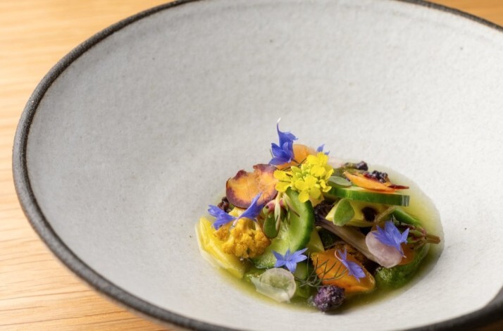 Seasonal vegetables and edible blooms arranged in a light herb broth, served in a dark-rimmed ceramic bowl at Jacaranda.