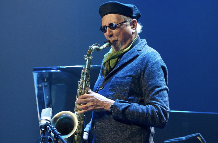 Musician Charles Lloyd playing a saxophone against a solid blue background.