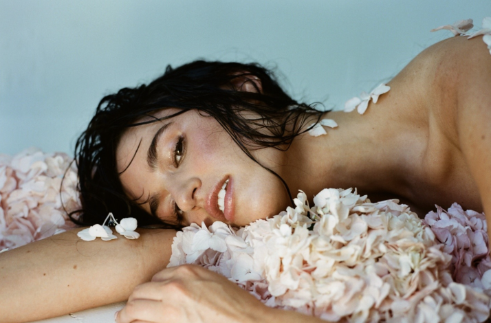 Singer Rozzi lying with wet black hair amidst pale pink flowers against a light blue background.