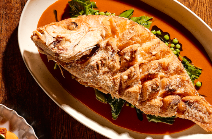 Salt-crusted whole roasted fish plated over vibrant red sauce with snap peas and greens, served at Little Water.