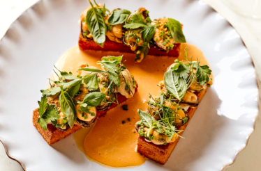 Three toasted brioche pieces topped with clams, fresh herbs, and microgreens in a golden sauce on a scalloped plate at Little Water.