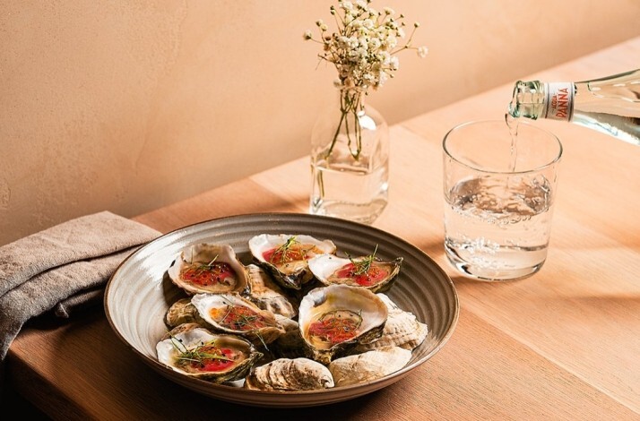Plate of oysters on the half shell with garnish on a wooden table, beside a glass of water being poured, served at River Twice.