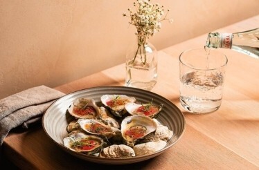 Plate of oysters on the half shell with garnish on a wooden table, beside a glass of water being poured, served at River Twice.