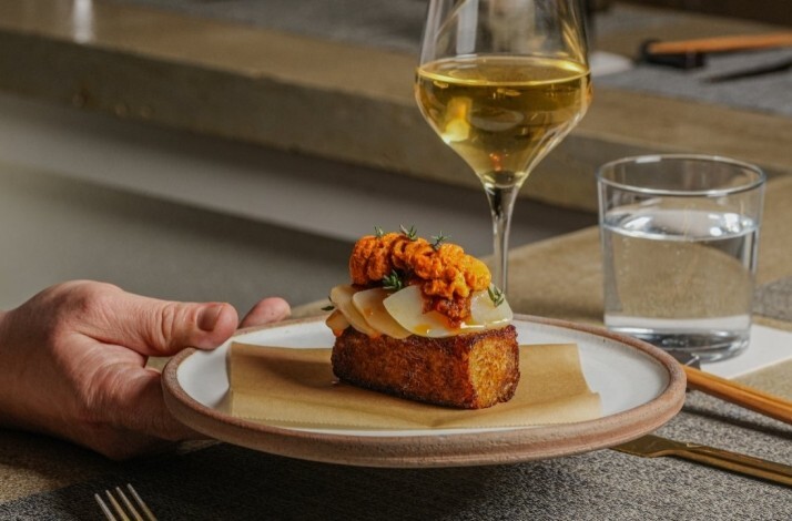 Hand holding a plated toast topped with sliced egg and orange roe, with a glass of white wine and water behind.