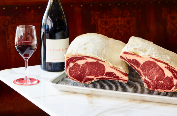 Dry-aged bone-in ribeyes displayed on a wire rack alongside a red wine bottle and glass at Ernesto's. 