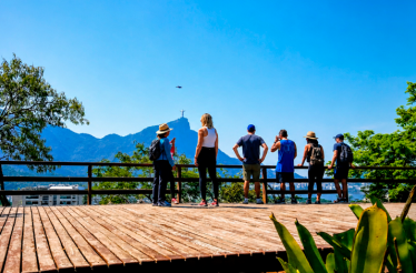 Visitors standing on a scenic observation deck overlooking Rio de Janeiro's mountains and city landscape under a clear blue sky.