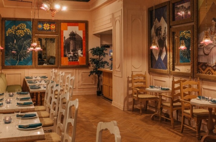 Restaurant interior featuring set dining tables, pendant lights, and colorful Indian-themed paintings on warm walls.
