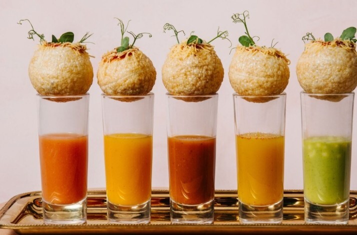 Row of five pani puri garnished with microgreens, each resting on a tall glass of flavored water in varying colors.