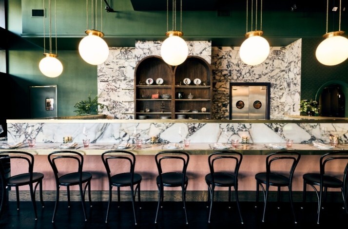 JouJou's marble bar counter lined with black bistro chairs, arched wood shelving, and globe pendant lights in a moody green-toned space.