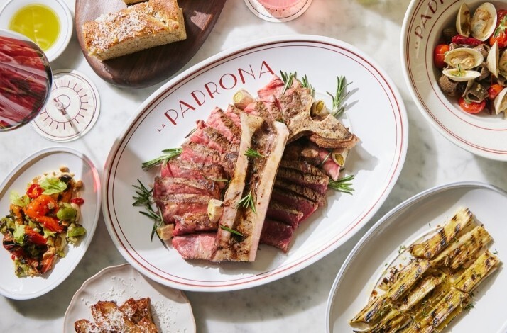 Sliced T-bone steak centred on a 'La Padrona'-branded oval platter, surrounded by clams, salad, focaccia, and grilled leeks.