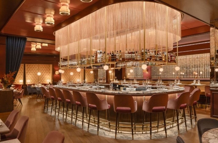 Oval bar at La Padrona Boston topped by a dramatic rose-gold fringe chandelier, surrounded by pink velvet bar stools.