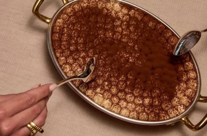 A large, oval-shaped serving dish filled with a creamy tiramisu dusted with cocoa powder.