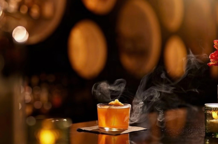 Smoked amber cocktail in a lowball glass on a bar top, with warm bokeh lights and floral accents in the background.