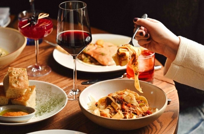 A hand lifting wide-ribbon pasta from a bowl surrounded by red wine, cocktails, focaccia, and shared plates at Porto Boston.