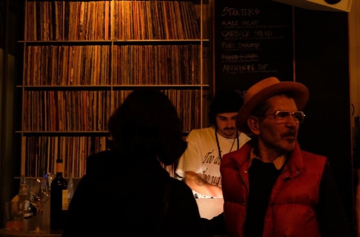 Dim bar with vinyl wall; people near counter, man in hat and red vest looks to the side.