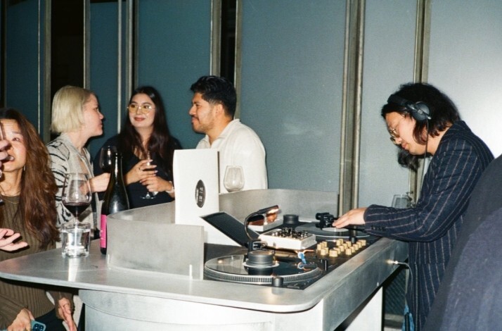 DJ mixes at a turntable while guests chat and drink wine at a small bar counter.