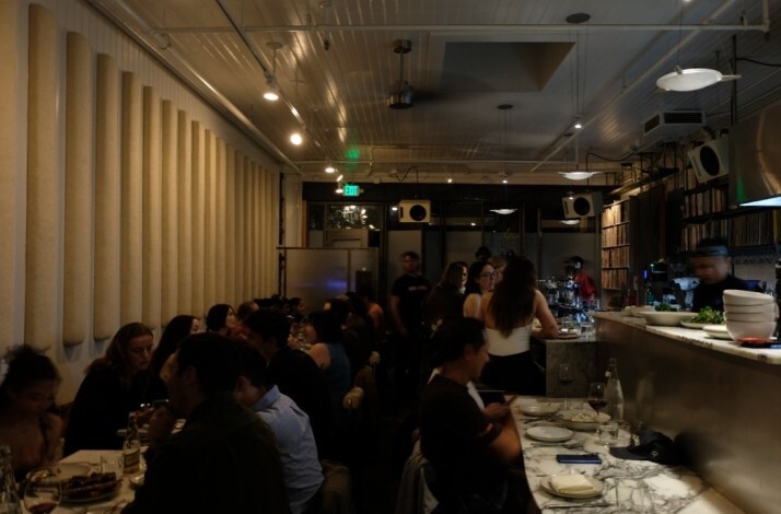 Busy restaurant dining room with long marble bar, seated guests, and warm overhead lighting.