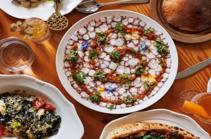 Overhead spread of dishes including sliced octopus carpaccio with harissa and borage flowers, black pasta and flatbread.