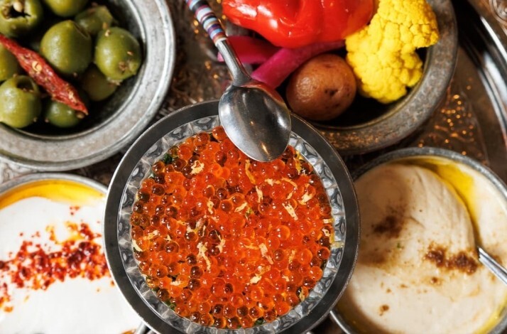 Overhead view of silver mezze bowls filled with salmon roe, green olives, hummus, and pickled vegetables.