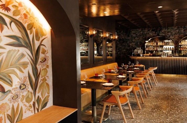 Moody dining room with floral wall mural, candlelit tables, and a curved back bar lined with bottles at Maria Isabel.