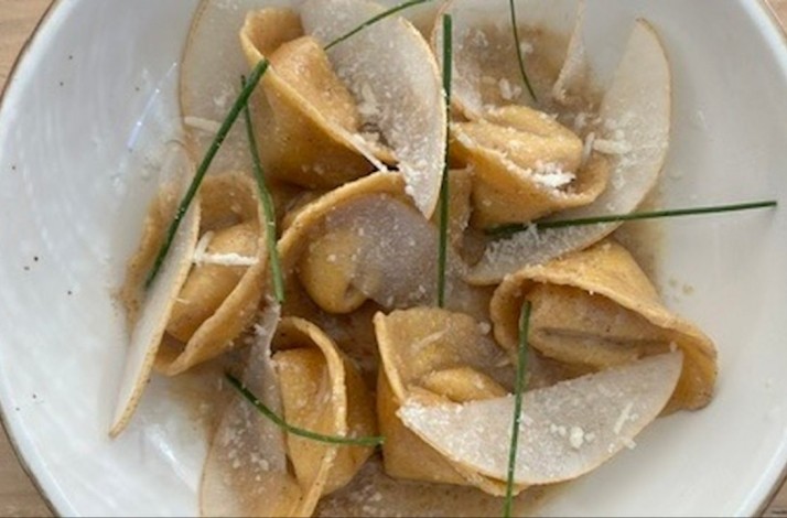Stuffed pasta parcels in golden broth, topped with shaved cheese and chive garnishes, served in a white ceramic bowl.