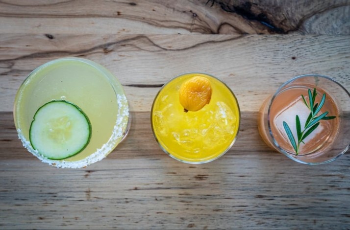 Trio of garnished cocktails on a wood surface, featuring cucumber, kumquat, and rosemary herb sprigs as finishing touches.