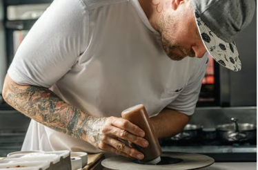 Chef Jeremy Ford focused on plating a dish in a restaurant kitchen while wearing a white apron and cap.