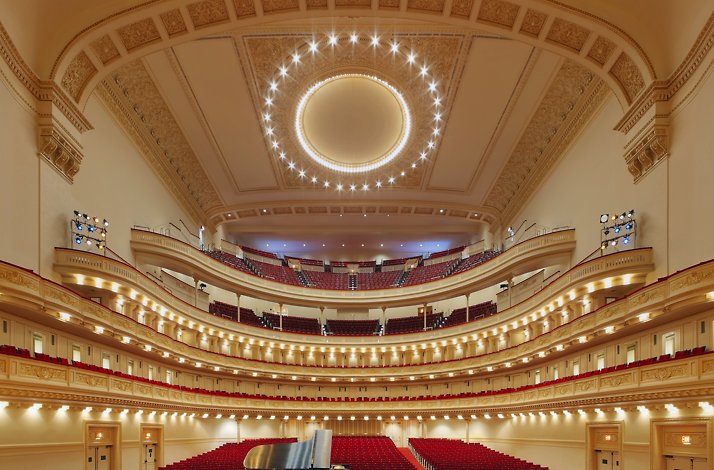 A grand, ornate concert hall interior of Carnegie Hall, surrounded by multiple tiers of red seating.