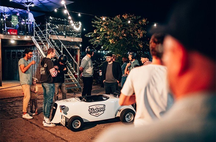 A group of men gathered around a HotRod car under the glow of fairy lights at night.