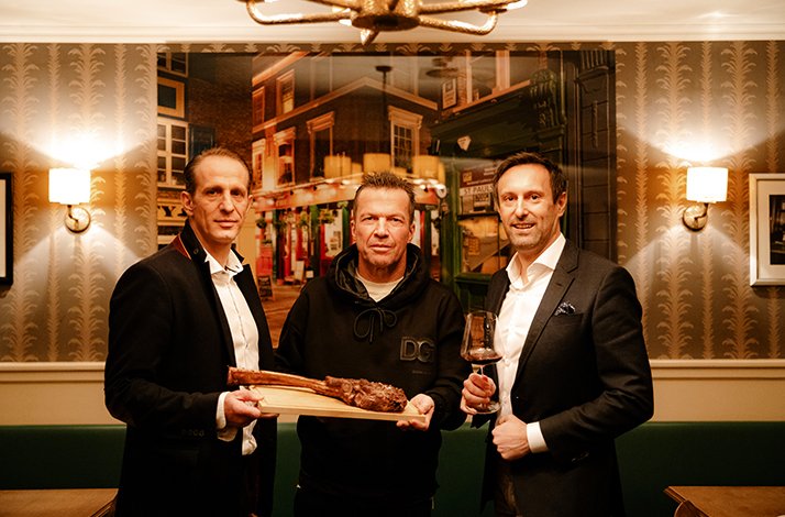 German football legend Lothar Matthäus posing with two other men holding a Tomahawk Steak and a wine glass.