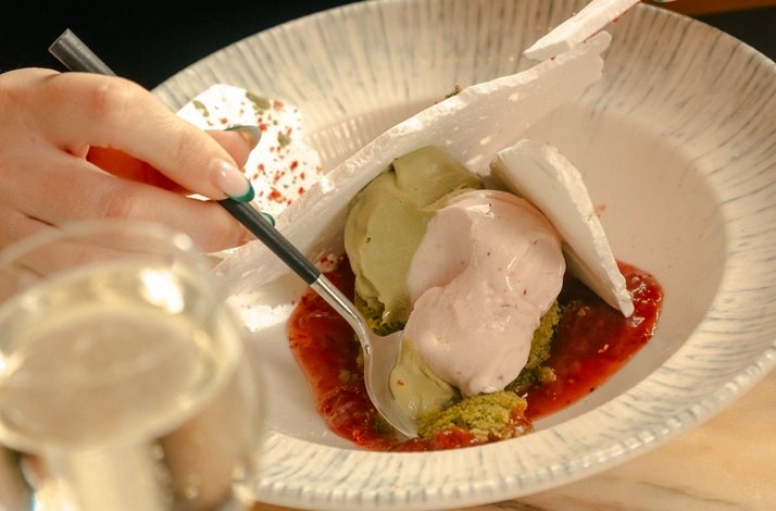 a plated dessert featuring green and pink ice cream, meringue shards, and a vibrant red fruit sauce, with a hand holding a spoon ready to take a bite at Jean-Georges at The Leinster.