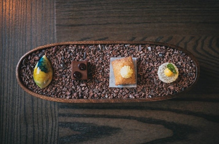 An elegant aerial view of four artfully crafted petit fours and confections presented on a rustic oval wooden tray filled with cacao nibs at Amai by Viktor, Dublin — featuring a glazed chocolate bonbon, a delicate pastry topped with cream, and two jewel-like sugar-coated treats.