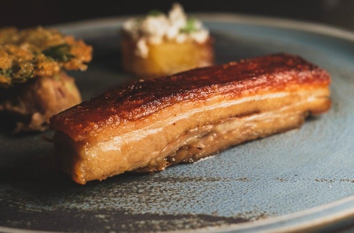 A beautifully caramelised and perfectly crisped slab of pork belly with layers of tender meat, plated on a speckled blue ceramic dish alongside herb-crusted and potato accompaniments, as part of the six-course tasting menu at Amai by Viktor, Michelin Guide-featured restaurant in Dublin.