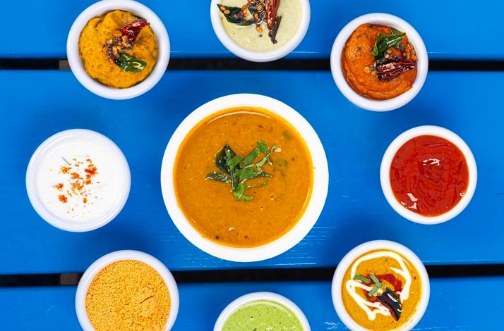 Colorful array of South Indian chutneys and curry served in small white bowls on a blue wooden table.