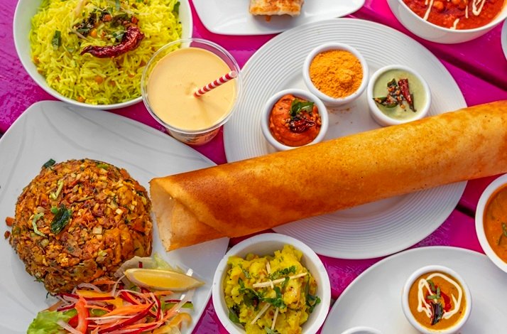 South Indian feast featuring a long crispy dosa, yellow rice, curries, and a mango lassi on a pink table.