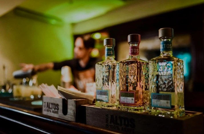 Three bottles of Altos tequila displayed on a bar counter with a bartender in the background. 