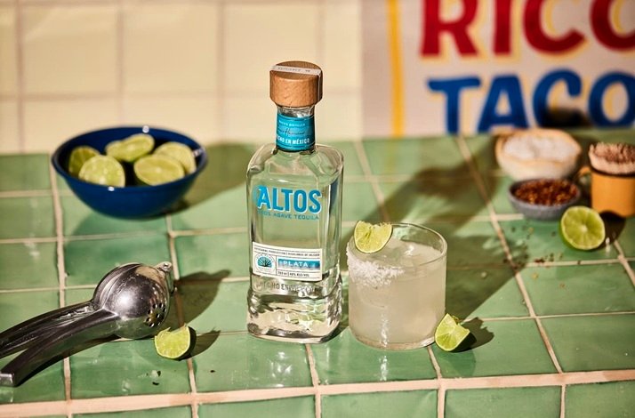 Altos Plata 100% Agave Tequila bottle beside a salt-rimmed margarita glass on green tiles, with limes and spices nearby.
