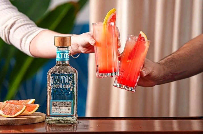 Two hands hitting glasses of paloma cocktail garnished with grapefruit beside an Altos 100% Agave tequila bottle.