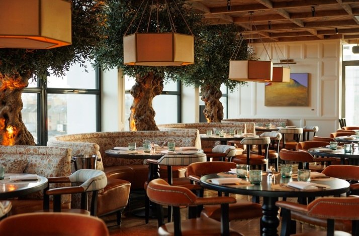 The elegant dining room of Jean-Georges at The Leinster, with round tables, plush seating, large windows, and decorative indoor trees creating a warm, inviting atmosphere.