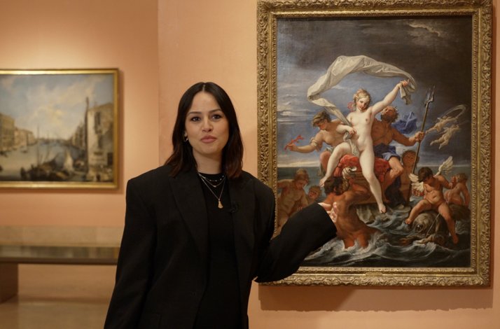 Photographer Rocío Aguirre standing in an art gallery, positioned between two framed oil paintings.
