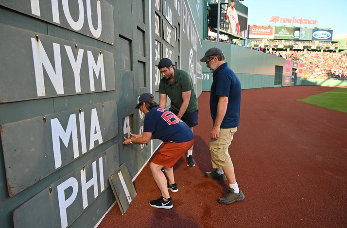 Three men are writing on a sign while attending a baseball game, focused on their task amidst the crowd.